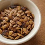 Cinnamon Sugar Roasted Pumpkin Seeds piled in a cream bowl with crisp amber edges and visible cinnamon sugar crystals